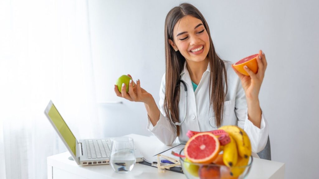 Profissional de Nutrição avaliando alimentos. Em sua mesa, há uma tigela com frutas, uma fita métrica corporal, um computador e um jarro de água.