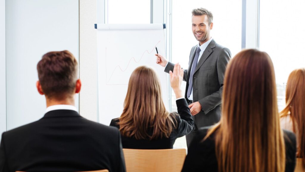 Processo de Treinamento e Desenvolvimento de colaboradores em uma organização. Na imagem, um profissional leciona uma aula para funcionários, que acompanham e questionam.