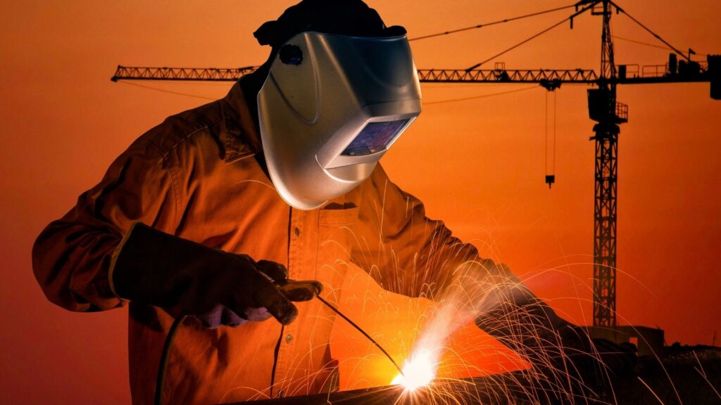 Trabalhador com máscara de proteção e luvas realizando processo de soldagem em estrutura metálica, com faíscas visíveis, ao ar livre durante o pôr do sol e guindastes ao fundo.