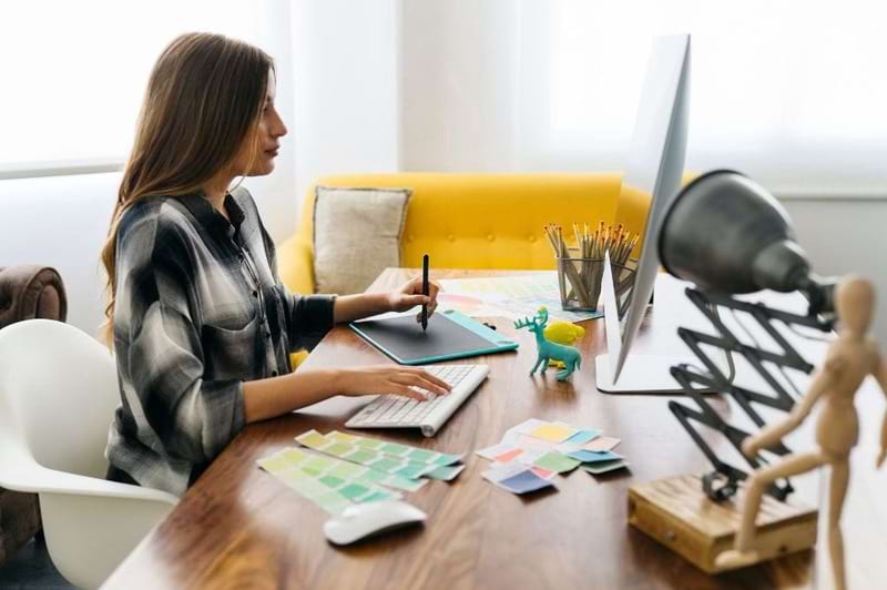 Estudante da faculdade de design gráfico trabalhando em seu computador, usando uma mesa de trabalho com materiais de design, como paletas de cores e um tablet gráfico. O ambiente é moderno, com iluminação suave e decoração minimalista, incluindo uma cadeira confortável e uma lâmpada de mesa.
