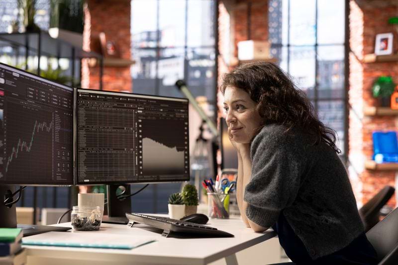 Profissional em ambiente de trabalho observando gráficos e dados em múltiplos monitores, exemplificando o trabalho de Análise e Desenvolvimento de Sistemas, com foco em análise de dados e monitoramento de desempenho de sistemas.