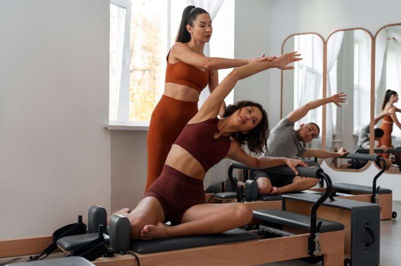 Instrutora de pilates orientando uma aluna em um exercício no reformer, com dois outros participantes ao fundo, em um ambiente moderno de estúdio de pilates.