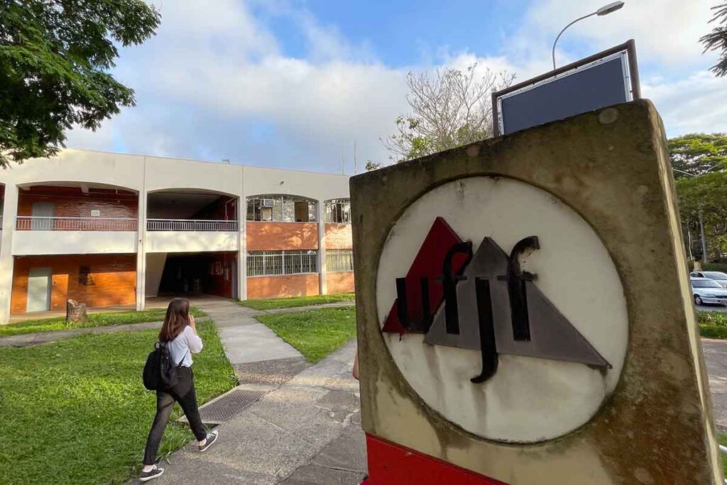 Estudante caminha pelo campus da UFJF próximo a placa com o logotipo da universidade, relacionada à lista de espera da UFJF.