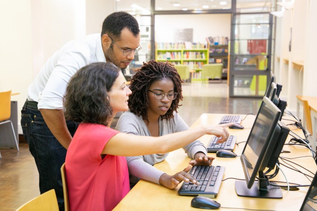Estudantes de ciência da computação com professor analisando atividades em computadores durante aula prática em laboratório de informática.