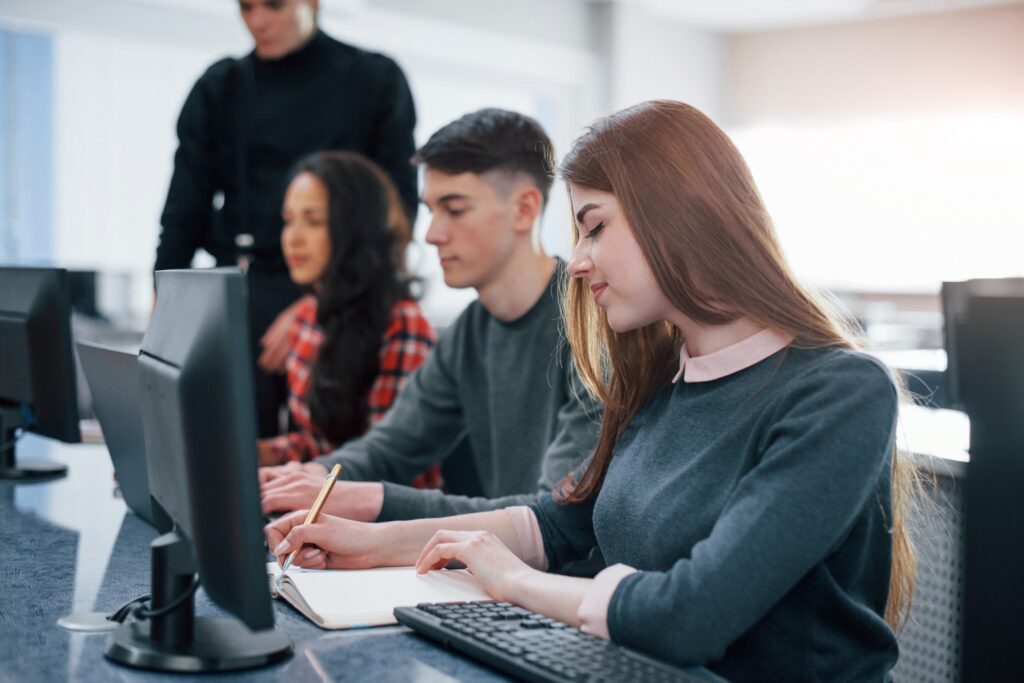 Estudantes de ciência da computação em laboratório de informática durante aula prática com computadores e professor orientando atividades.