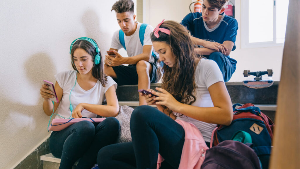 Adolescentes mexendo em celular sentados na escada da escola; conheça as principais gírias da geração Z.