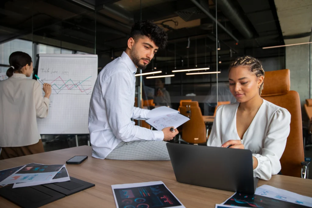 Profissionais de gestão empresarial, juntos, conversando. A mulher está sentada, mexendo no notebook, enquanto o homem está apoiado na mesa.