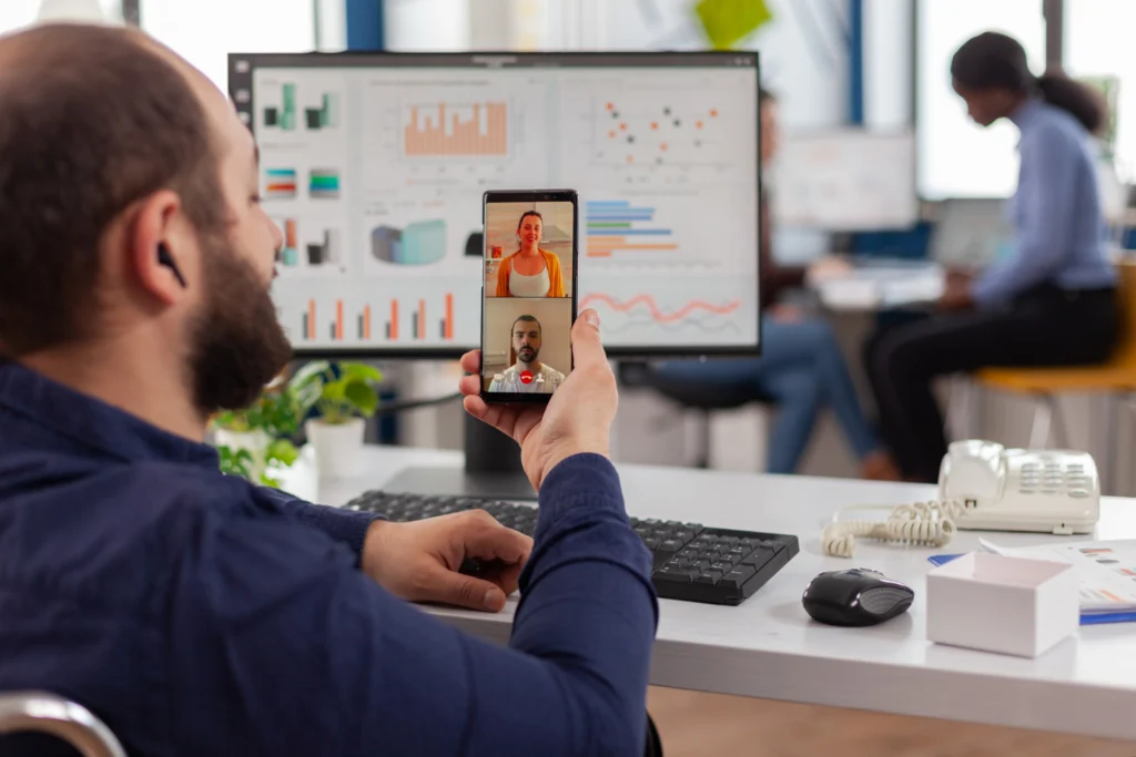 Homem com pós em comunicação e marketing atuando na área, em um escritório. Ele conversa com duas pessoas do time através de videochamada no celular. Na sua frente, tem uma mesa com computador.