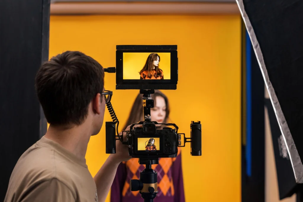 Homem com pós em direção de arte conduzindo uma câmera, de frente a uma mulher que está posando em um fundo amarelo.