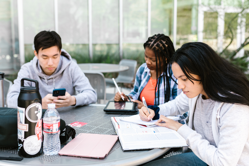 três adolescente estudando na mesa com materiais de estudo, cadernos e dispositivos digitais