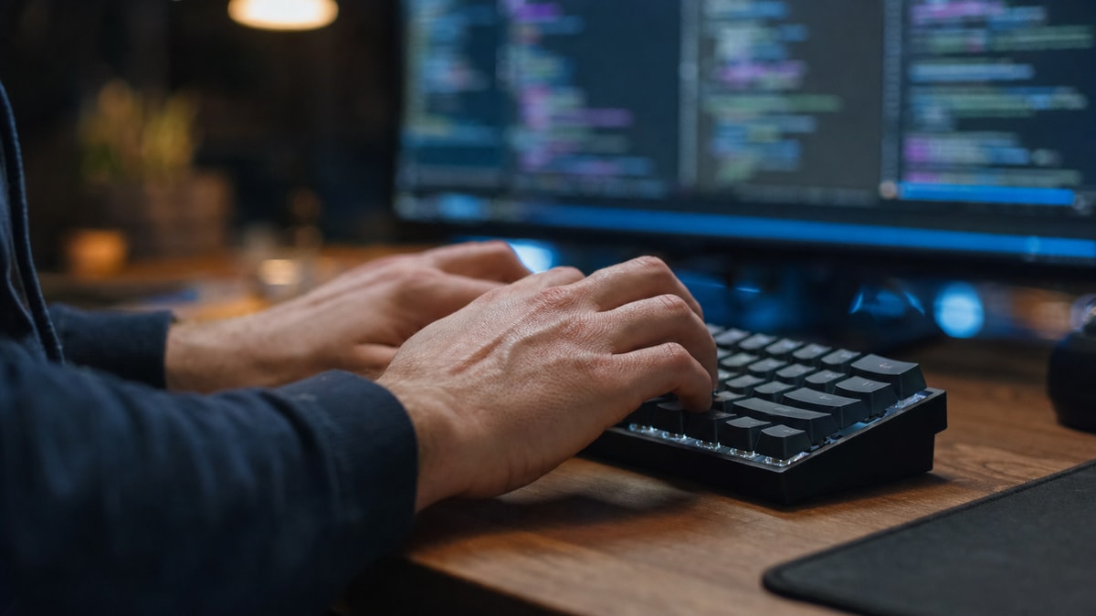 Mãos digitando em teclado mecânico com monitor ao fundo apresentando painéis organizados de desenvolvimento.