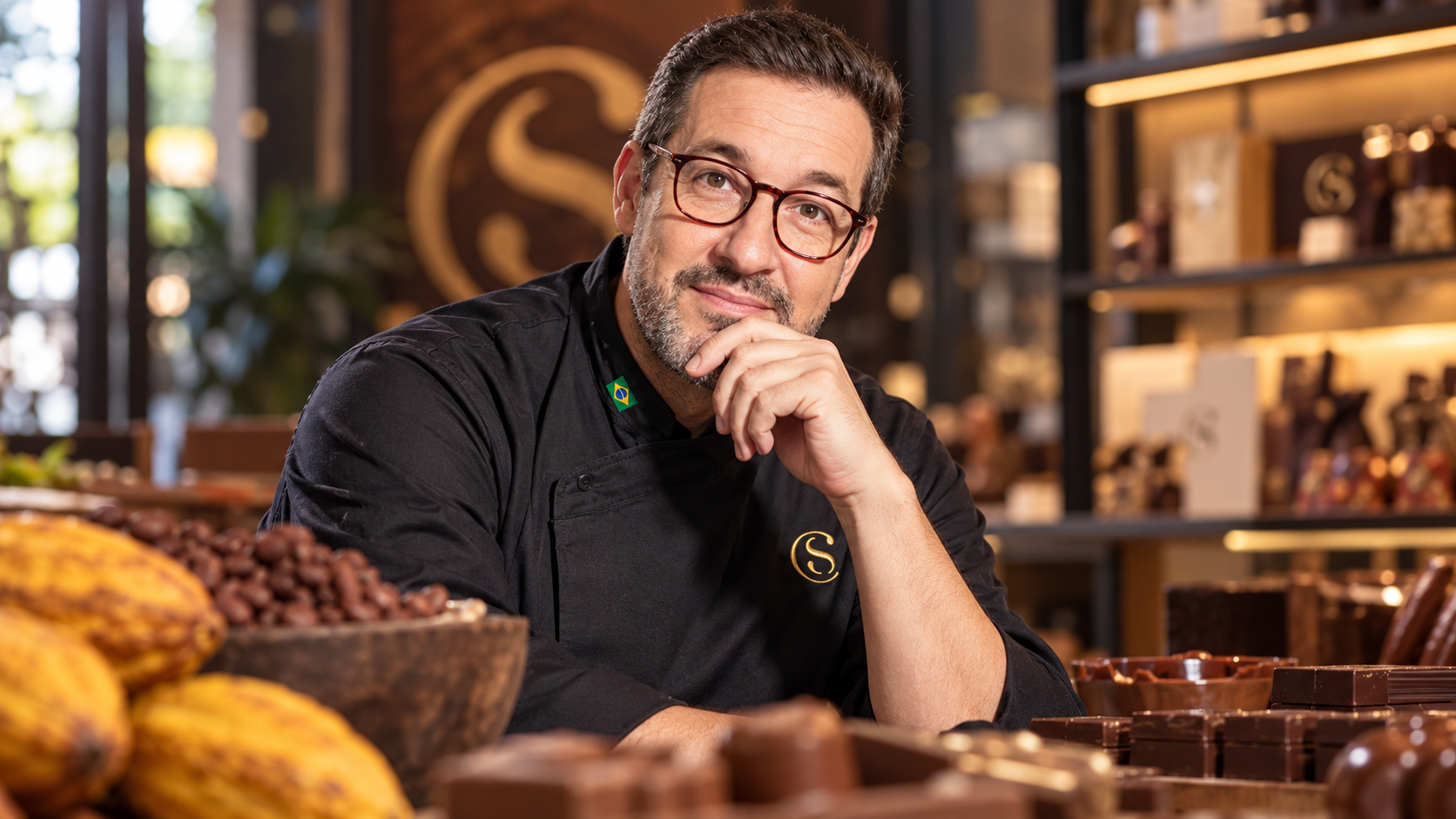 Retrato de Alê Costa, de dólmã preto com o logotipo da Cacau Show, entre frutos de cacau e chocolates