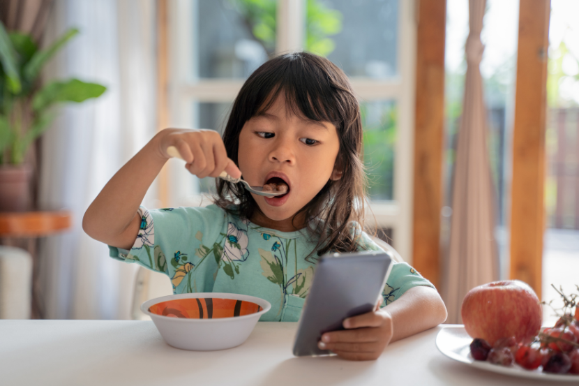 criança pequena mexendo no celular enquanto come na mesa